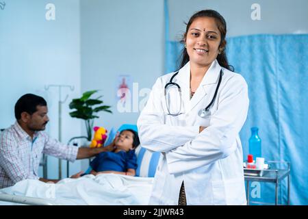 Glücklicher, selbstbewusster Arzt mit gekreuzten Armen, der vor dem Patienten steht, indem er die Kamera im Krankenhaus betrachtet - Konzept der medizinischen Behandlung, professionell Stockfoto