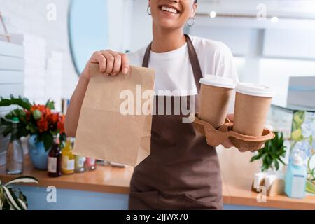 Beschnittene Ansicht des afroamerikanischen Verkäufers, der Papiertüte und Kaffee zum Mitnehmen in Süßwaren hält Stockfoto