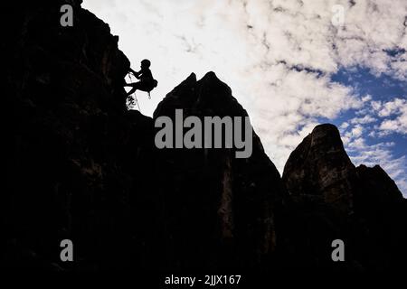 Flager Winkel voller Länge der Silhouette einer aktiven, nicht erkennbaren Frau, die auf einer felsigen Klippe gegen den Himmel klettert Stockfoto