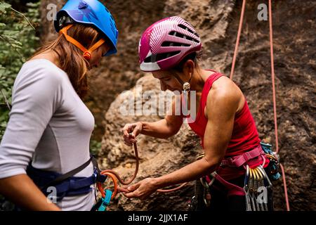 Aktive ältere Frau, die einer Freundin beim Seilbinden am Klettergurt hilft, während sie sich auf das Klettern vorbereitet Stockfoto