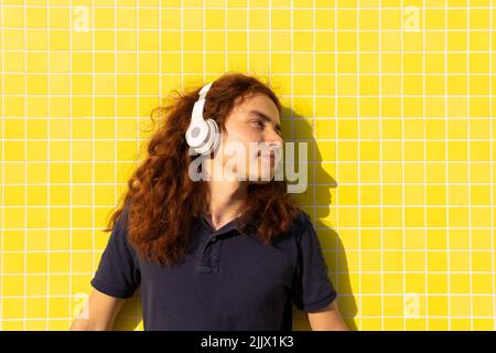 Junge Frau mit Kopfhörern schaut weg, während sie gegen die gelbe Wand steht Stockfoto
