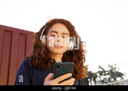 Von unten junge Frau mit Kopfhörern, die auf dem Smartphone gegen den Himmel stöbert Stockfoto