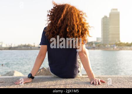 Rückansicht einer jungen Frau mit Rotschopf, die in der Stadt am Strand sitzt Stockfoto