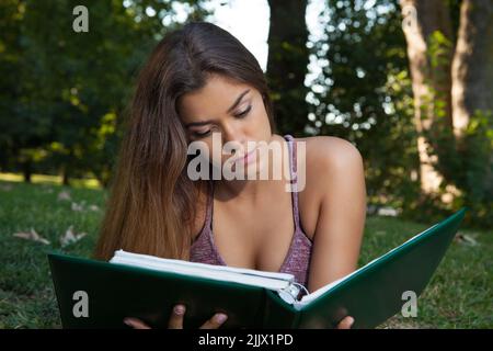 Eine junge hispanische Frau, die ein Notizbuch liest und in einem Park studiert Stockfoto