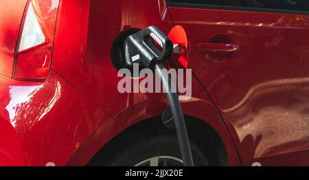 Gaspistole im Tank aus rotem Auto, das während des Betankens auf der Straße mit hellem Sonnenlicht in der Stadt auf der Tankstelle geparkt ist Stockfoto