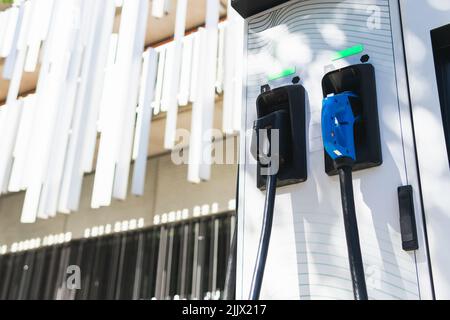 Moderne öffentliche Ladestation für Elektrofahrzeuge mit Schusswaffen in der Nähe eines modernen mehrstöckigen Gebäudes auf der Straße mit Sonnenlicht in der Stadt Stockfoto