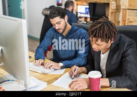 Konzentrierte, multirassische männliche Mitarbeiter, die am Computer tippen und Notizen auf Dokumente schreiben, während sie gemeinsam an einem Geschäftsprojekt im Büro arbeiten Stockfoto