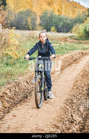 Der blonde Junge im lässigen Outfit fährt auf der Landstraße an Bäumen mit vergilbten Blättern vorbei auf dem Fahrrad. Schuljunge genießt sportliche Aktivitäten am Herbsttag Stockfoto