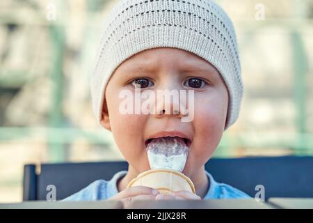 Kleinkind Junge isst Eis mit Vergnügen und schaut auf Kamera Nahaufnahme. Junge in weiß gestricktem Hut leckt Eis auf der Bank auf dem Spielplatz sitzen. Gehen Sie hinein Stockfoto