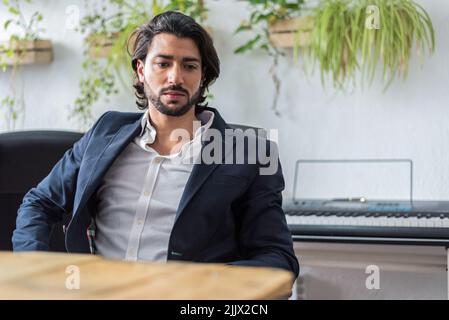 Selbstbewusster männlicher Manager mit Perlen in formellem Anzug, während er während der Arbeit am Tisch in einem hellen, modernen Büro mit grünen Pflanzen sitzt Stockfoto
