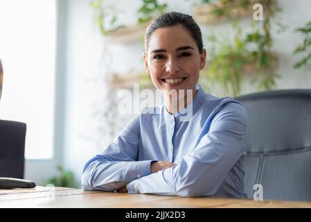 Fröhliche Managerin aus Kaukasien in weißer Bluse mit lockigen Haaren lächelnd und Blick auf die Kamera, während sie hinter dem Tisch im sonnendurchfluteten Büro sitzt Stockfoto