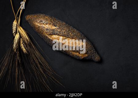 Draufsicht frisch gebackenes Baguette, bedeckt mit Mohn, in der Nähe von einem Haufen Weizenspikes auf schwarzem Hintergrund im Studio platziert Stockfoto