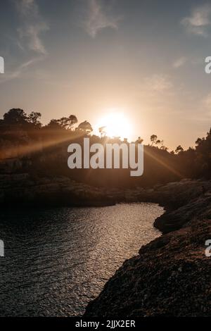 Helles Sonnenlicht, das über einer malerischen Bucht mit felsigen Klippen scheint, die das plätschernde Meer umgeben, und den wolkigen Himmel auf den Balearen Stockfoto