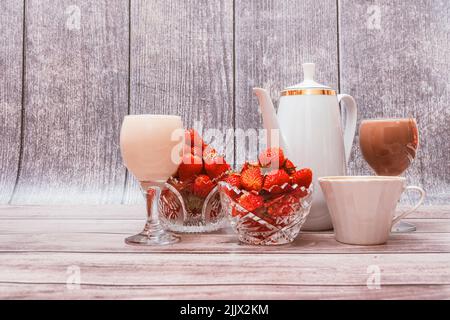 Zwei Vasen mit reifen Erdbeeren, einer weißen Kaffeeschale, einer Tasse Kaffee und zwei Gläsern Cocktails stehen auf Holzhintergrund. Stockfoto