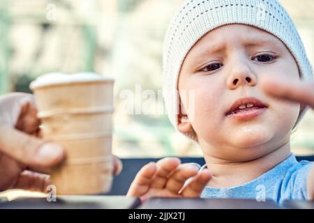 Papa füttert fokussiertes Kleinkind mit Eis auf der Spielbank. Junge in weiß gestrickter Mütze sitzt am Tisch und schaut mit Interesse aus der Nähe zur Seite Stockfoto