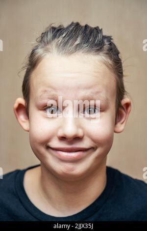 Netter Schuljunge mit blonden Haaren und dunkelbraunen Augen lächelt weit in die Kamera. Porträt eines Jungen im schwarzen T-Shirt gegen beige Wand Nahaufnahme Stockfoto