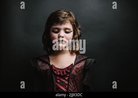 Scary Mädchen in Kleid mit schrecklichen dunklen Eyeliner und Lippenstift Blick nach unten, während auf schwarzem Hintergrund während der Halloween-Feiertagsfeier stehen Stockfoto