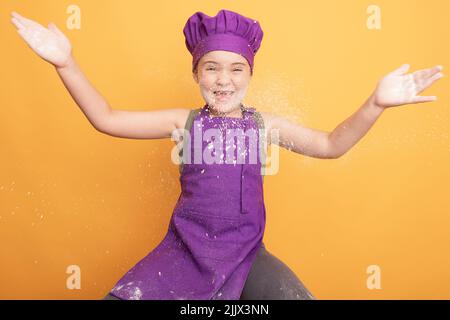 Optimistisches Mädchen in lila Chefuniform klatscht Hände mit weißem Mehl, während sie auf gelbem Hintergrund im modernen hellen Studio sitzt und den Kamerafelder anschaut Stockfoto