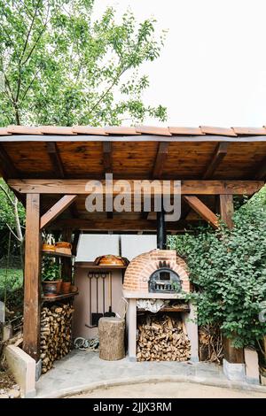 Traditioneller rustikaler Ofen mit Haufen gehacktem Brennholz in Holzpavillon im Hinterhof des ländlichen Hauses inmitten grüner Bäume Stockfoto