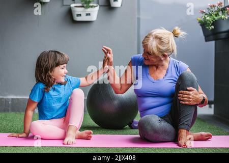 Lächelndes Mädchen, das der Großmutter, die auf einer Matte im Fitnessclub sitzt, fünf hoch gibt und sich anschaut Stockfoto