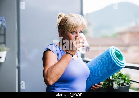 Fröhliche ältere Frau, die eine gerollte Yogamatte mit einem Glas Wasser nach dem Training hält und wegschaut Stockfoto