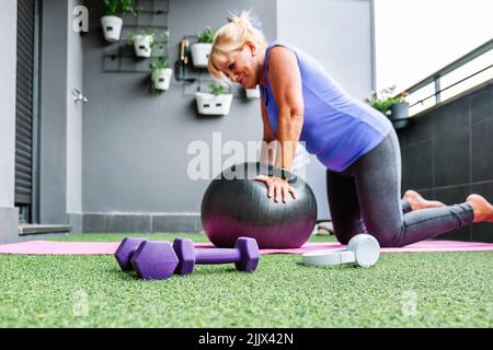 Ganzkörper-Seitenansicht einer älteren Frau, die mit dem Fitnessball auf dem Balkon trainiert Stockfoto