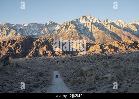 Rotes Wohnmobil, das am Wintertag in Alabama Hills, Califo, zwischen Feldern auf einer langen Straße gegen eine Reihe rauer, mit Schnee bedeckter Felsklippen fährt Stockfoto