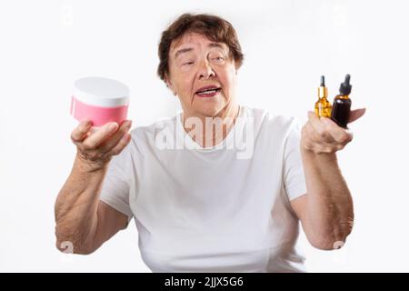 Porträt einer älteren lächelnden Frau, die zwei Flaschen Kosmetik und ein Glas Creme in den Händen hält. Weißer Hintergrund. Hautpflege und Gesundheitskonzept Stockfoto