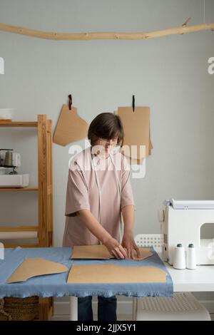 Fokussierter professioneller Schneidermeister mit Maßband, der Muster auf Stoff macht, während er am Tisch mit spezieller Ausrüstung in einem hellen Werkstatelei arbeitet Stockfoto