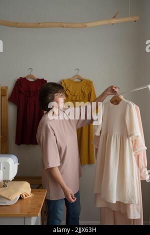 Frau mittleren Alters in Gläsern, die handgemachtes Kleid vorführt, während sie in der Nähe der Nähmaschine im Werkstatter steht Stockfoto