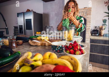Frau, die grünen Grünkohl mit gehackter Ananas in eine Mixschüssel gibt, während sie am Tisch Smoothie mit verschiedenen Früchten und Beeren zubereitet Stockfoto