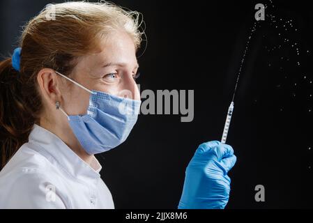 Der Arzt sprüht aus einer Spritze auf einen schwarzen Hintergrund. Der Arzt lächelt unter seiner Schutzmaske, die Droge aus der Spritze ergießt sich in die Luft Stockfoto