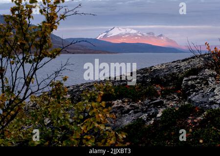 Episches Sonnenaufgangslicht beleuchtet felsigen, gletscherbedeckten Gipfel mit dunklem bewölktem Himmel im Hintergrund. Ahkka Peak im wunderschönen goldenen Stundenlicht, Stora Stockfoto