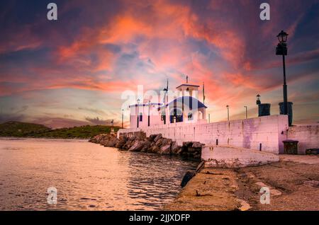 Kleine griechische Kapelle mit weißen Dächern auf Klippen über dem Meer und einer kleinen Bucht unter einem dramatischen Himmel bei Sonnenuntergang Stockfoto