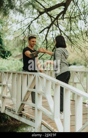 Nettes Paar, das sich die Hände schüttelt und lacht, während es auf der weißen Brücke im Park steht Stockfoto