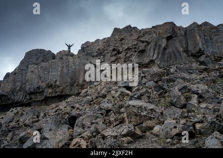 Dettifoss, Island - 06.15.2017: Mann mit ausgestrecktem Arm, der auf einem schwarzen Basaltgestein in der Nähe des Dettifoss-Wasserfalls in Island steht. Stockfoto