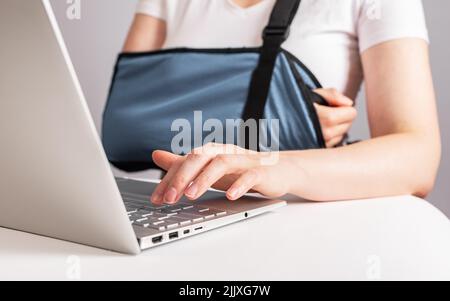 Frau mit schmerzhaftem Arm in einem Tragetuch, die am Laptop arbeitet. Die Frau trägt einen Verband, um den verletzten Ellbogen, die Schulter, den Unterarm oder das Handgelenk zu stützen, während sie mit dem Computer am Tisch sitzt. Hochwertige Fotos Stockfoto