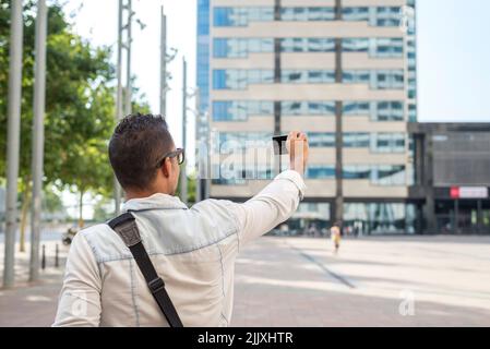 Junger Mann mit Sonnenbrille und einer Schultertasche, der mit einem Telefon in der Stadt Fotos gemacht hat Stockfoto