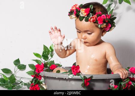 Nahaufnahme eines wunderschönen braunen Latina-Mädchens, das spielt, während es in einem grauen Eimer gebadet wird. Das 9 Monate alte Mädchen spritzt Wasser mit ihrem ha Stockfoto