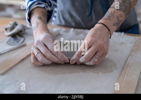 Töpferin, die am Tisch steht und mit Ton arbeitet, weibliche Hände, die im Atelier handgemachte Töpferwaren anfertigten Stockfoto