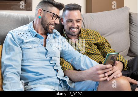 Glückliches junges schwules Paar, das im Wohnzimmer auf einem Sofa sitzt und das Mobiltelefon benutzt. Hochwertige Fotografie. Stockfoto