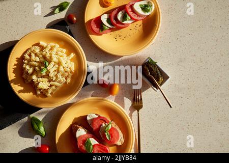 Italienische Pasta-Fusilli, Caprese-Salat - Tomaten, Basilikum, Mozzarella und Pesto. Brotvorspeise mit Caprese-Salat. Draufsicht. Harte Schatten. Italienisches Abendessen Stockfoto