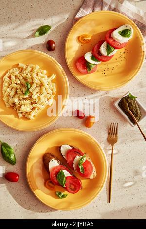 Mediterrane Nudelfusilli, italienischer Caprese-Salat - Tomaten, Mozzarella, Basilikum und Pesto. Brotvorspeise mit Caprese-Salat auf orangen Tellern unter trendigen Schatten. Draufsicht Essen flach legen Stockfoto