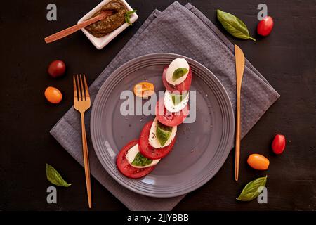 Italienischer mediterraner klassischer Caprese-Salat mit Mozzarella, Tomaten, Basilikum und Pesto-Sauce auf schwarzem rustikalem Hintergrund. Essen von oben. Stockfoto