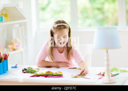 Kind schafft Bild mit bunten Blättern. Kunst und Kunsthandwerk für Kinder. Kleines Mädchen macht Collage Bild mit Regenbogenpflanzen Blatt. Stockfoto