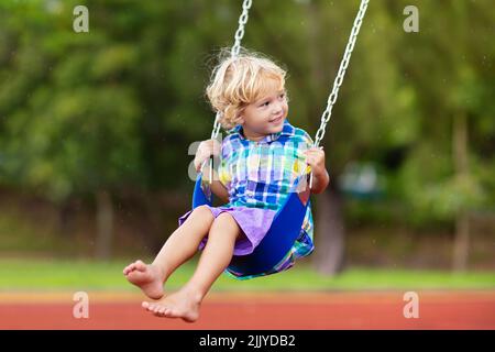 Kinder spielen auf dem Spielplatz im Freien bei Regen. Kinder spielen auf der Schule oder im Kindergarten. Aktives Kind auf farbenfroher Schaukel. Stockfoto