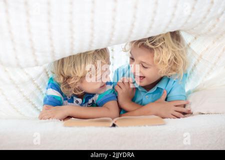 Lesebuch für Kinder im Bett unter der Strickdecke. Zwei Brüder spielen zusammen. Kinder gemütliches Zimmer im Hyggge-Stil. Kleiner Junge, der Hausaufgaben macht, bevor er schläft. Stockfoto