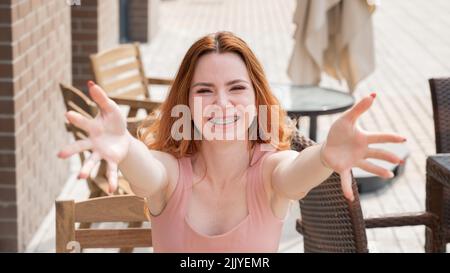 Die junge rothaarige Frau sitzt in einem Straßencafé und streckt ihre Arme zur Umarmung. Stockfoto