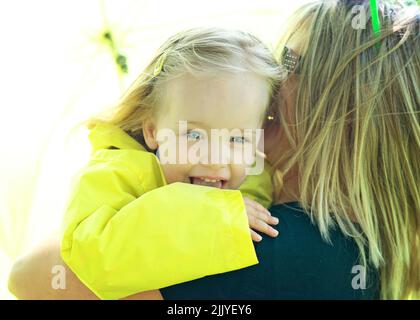 Kleines blondes Mädchen, das lachend über Mutters Schulter blickt. Stockfoto