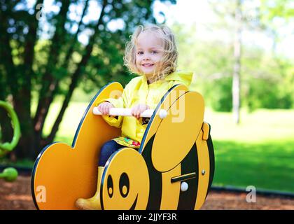 Blonde Kleinkind Mädchen in gelbem Regenmantel auf Bienenritt, Spielplatz. Stockfoto